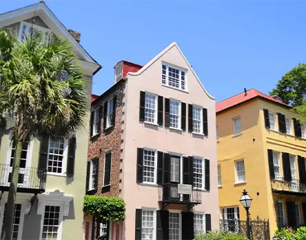 A row of colorful historic three-story homes in Charleston, South Carolina, featuring pastel pink, yellow, and green exteriors with traditional black shutters and wrought iron balconies under a clear blue sky.